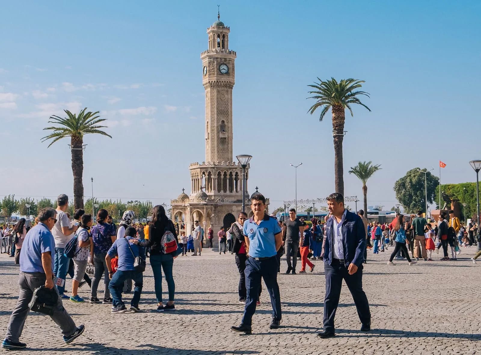Часовая башня Измира (İzmir Saat Kulesi) возвышается над площадью Конак в окружении пальм и голубей, на фоне Измирского залива
